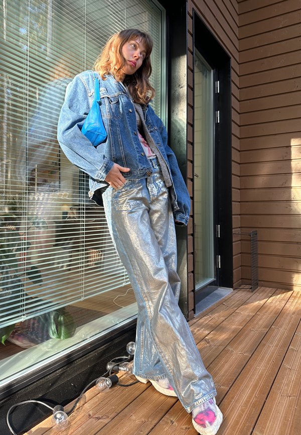 Denim jakna preko roza majice, v kombinaciji z bleščečimi srebrnimi širokimi hlačami. Majhna modra torbica in pisanih nogavicah dopolnjujeta outfit. Ozadje iz lesenega poda.