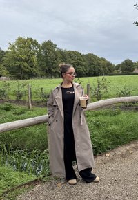 Beige trenchcoat över en svart topp med urklipp, parat med svarta vida byxor och ljusa tan slip-on skor. Håller en kaffekopp.