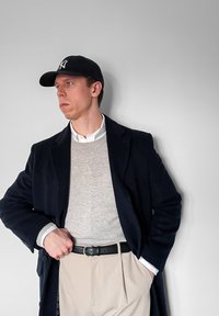 Homme portant une casquette de baseball noire, un manteau noir, un pull gris, une chemise blanche et un pantalon beige, debout avec une main dans la poche contre un mur blanc.
