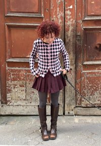 Παιδί με σγουρά κόκκινα μαλλιά φοράει πουλόβερ με σχέδιο, πλισέ φούστα, γκρίζες καλσόν και καφέ μπότες, στέκεται μπροστά σε φθαρμένες ξύλινες πόρτες.