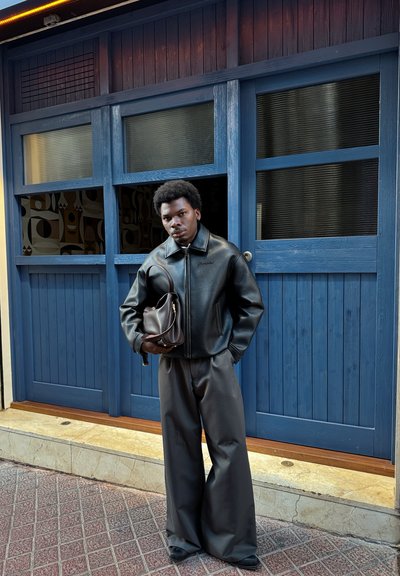 Chaqueta de cuero negra con cuello alzado, pantalones grises de pierna ancha y un bolso marrón. Puerta de madera azul con paneles de vidrio en el fondo.
