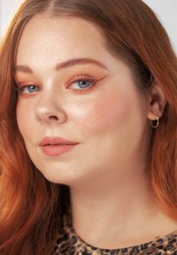 Ung kvinde med rødt hår, blå øjne, vingeformet kobberøjenskygge, nude læbestift, næsering og ørering i ring, iført en leopardprint top.