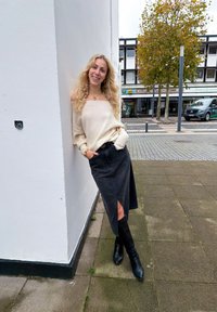 Suéter off-shoulder tricotado em bege, saia de ganga escura com racha lateral, botas pretas até ao joelho e colar de coração dourado. Ambiente urbano.