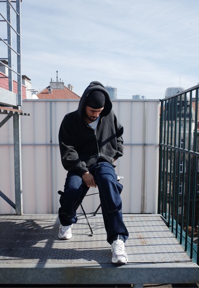 Sudadera negra con cremallera, jeans oscuros holgados y zapatillas blancas. Sentado en una silla plegable sobre una superficie metálica con un fondo gris.