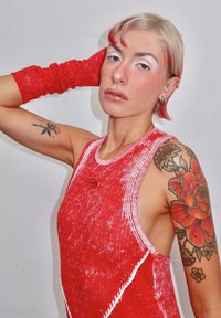 Jeune personne aux cheveux blonds courts avec des pointes rouges, portant un haut rouge sans manches et un gant en dentelle, montrant des tatouages floraux sur les deux bras.