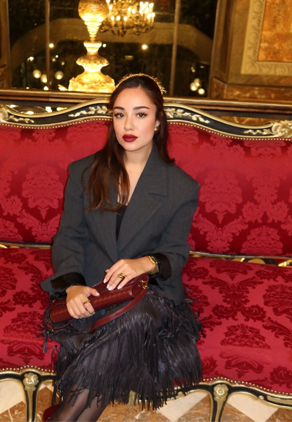 Donna seduta su un divano in velluto rosso. Indossa un blazer grigio oversize, una gonna nera con frange e tiene una borsa color bordeaux. Dettagli dorati visibili.