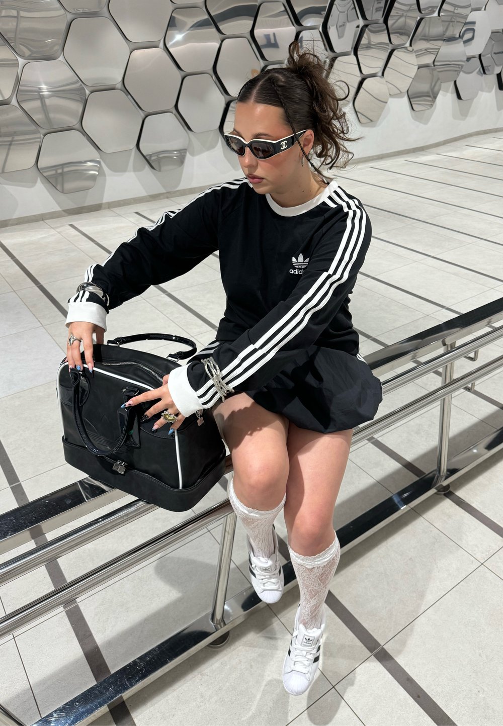 Camisa preta de manga longa com riscas brancas, saia preta, meias brancas até ao joelho e mala preta. Os acessórios incluem anéis e óculos de sol.