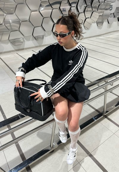 Camisa negra de manga larga con rayas blancas, falda negra, calcetas blancas hasta la rodilla y bolso negro. Los accesorios incluyen anillos y gafas de sol.