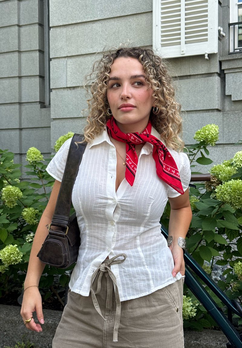 Frau mit lockigem Haar, die ein weisses Hemd mit Knöpfen, einen roten Schal, beige Hosen und eine Schultertasche trägt, steht draussen nahe grünen Pflanzen und einer grauen Steinmauer.
