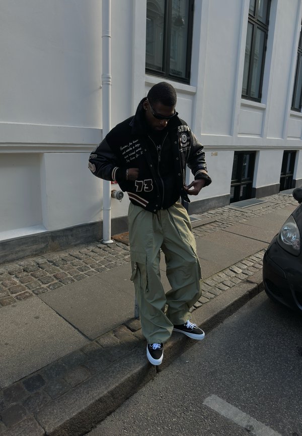 Mees, kellel on seljas must jakk ja khaki värvi cargo-püksid, seisab tänava ja valge hoone kõrval kõnniteel.