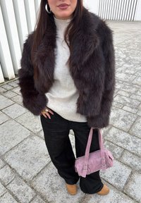 Femme portant une veste en fourrure marron foncé, un pull à col roulé beige, un pantalon noir, des bottines beige, des bagues en or, et tenant un sac à main en daim rose à l'extérieur.