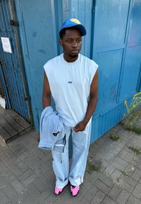 Gaiši zils bezpiedurkņu tops, vaļīgas gaiši zilas džinses, turēdams pelēku kapuci. Melns logo uz kapuces. Rozā apavi, zils cepure ar dzelteniem akcentiem.