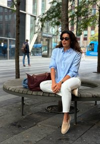 Chemise bleu clair à boutons, pantalon blanc, chaussures beige à enfiler ; assis sur un banc en bois circulaire avec un sac bordeaux et une veste bleue à proximité.