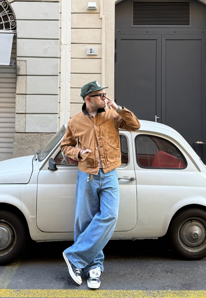 Man in brown jacket, blue jeans, and green cap leaning against small white vintage car on city street.
