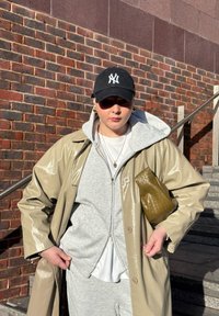 Personne portant un manteau beige brillant sur un sweat à capuche gris et un pantalon de survêtement, casquette noire, tenant une pochette tissée vert olive, debout près d'un mur en briques et des escaliers.