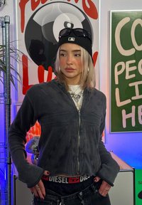 Chaqueta de cremallera gris oscuro con mangas largas, material texturizado, superpuesta sobre una camisa blanca, combinada con jeans negros y una gorra de béisbol.