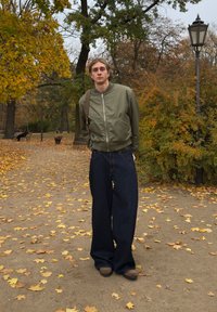 Roheline pommitaja jope, laiad tumedat sinist tooni teksad ja pruunid kingad. Modell seisab rajal, mida ümbritseb sügisesed lehed ja põõsastik.