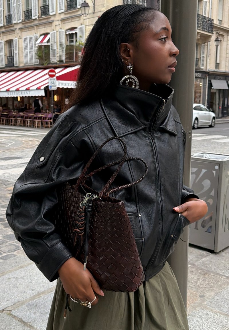Frau trägt eine schwarze Lederjacke und grosse Ohrringe, hält eine geflochtene dunkelbraune Handtasche und steht in einer Stadtstraße nahe einem Café.