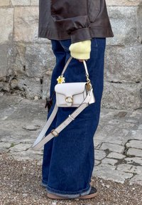 Small white leather crossbody bag featuring a gold circular clasp and floral accent, teamed with dark blue wide-leg jeans and a brown jacket.