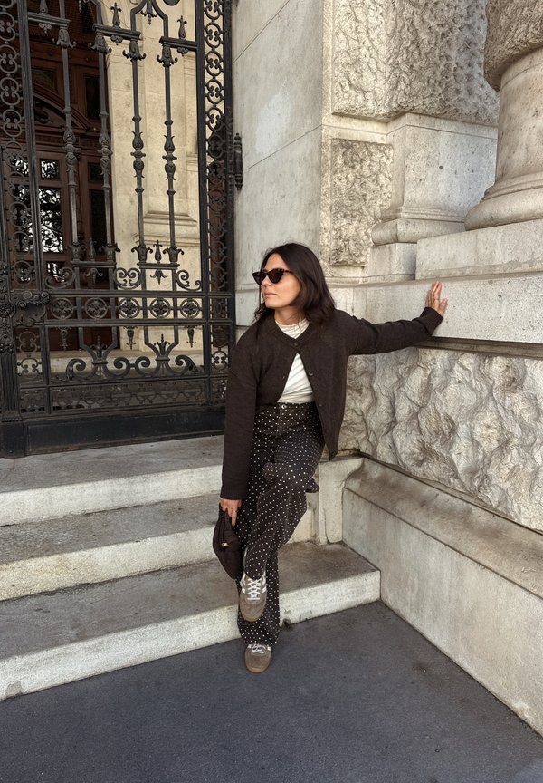 Mujer con gafas de sol y cárdigan oscuro se apoya en un muro de piedra junto a una puerta de hierro forjado, sosteniendo un sombrero y apoyando un pie en las escaleras.