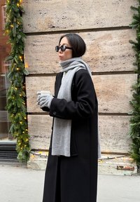 Sort frakke med store knapper, gråt strikket tørklæde og handsker. Står foran en tekstureret væg med grøn guirlande og lys.