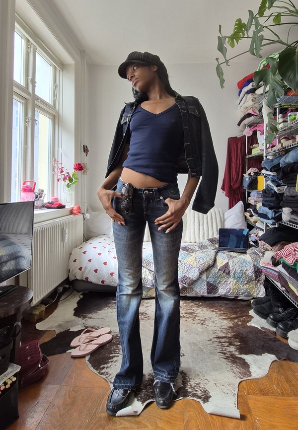 Personne portant un chapeau foncé, un haut bleu marine, une veste en jean et un jean évasé, debout dans une chambre près d'un lit, d'étagères, d'une plante d'intérieur et d'une fenêtre.