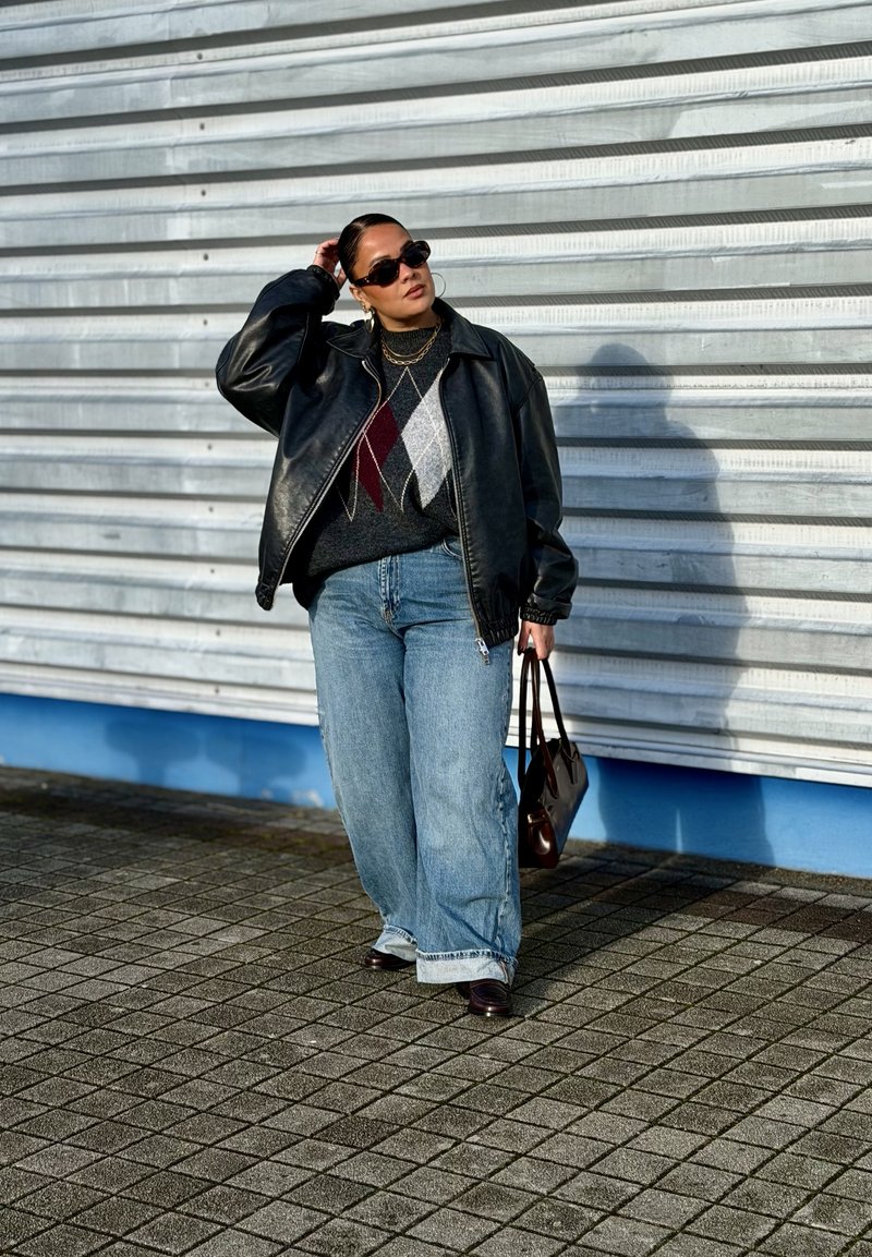 Woman in black leather jacket, argyle sweater, wide jeans, sunglasses, holding brown bag, standing on tiled pavement against metal shutter background.