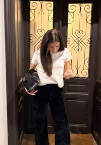 Bílé tričko s krátkým rukávem, černé sametové široké kalhoty, drží černou koženou tašku. V pozadí dekorativní dvojité dveře s kovanými železnými detaily.