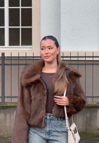 Giacca corta in ecopelliccia marrone con cappuccio, indossata sopra un top aderente e jeans in denim a vita alta. Gli accessori includono una piccola borsa.