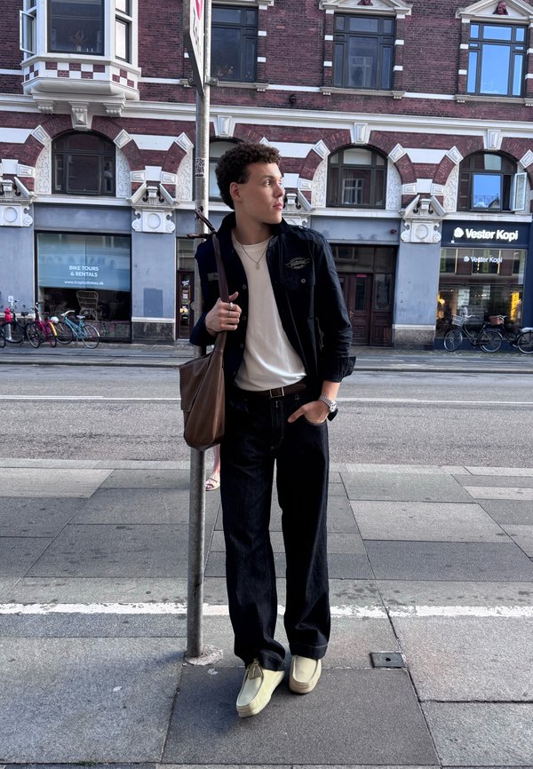 Giovane con capelli ricci che indossa giacca nera, camicia bianca, jeans scuri e scarpe beige, appoggiato a un palo su un marciapiede urbano.