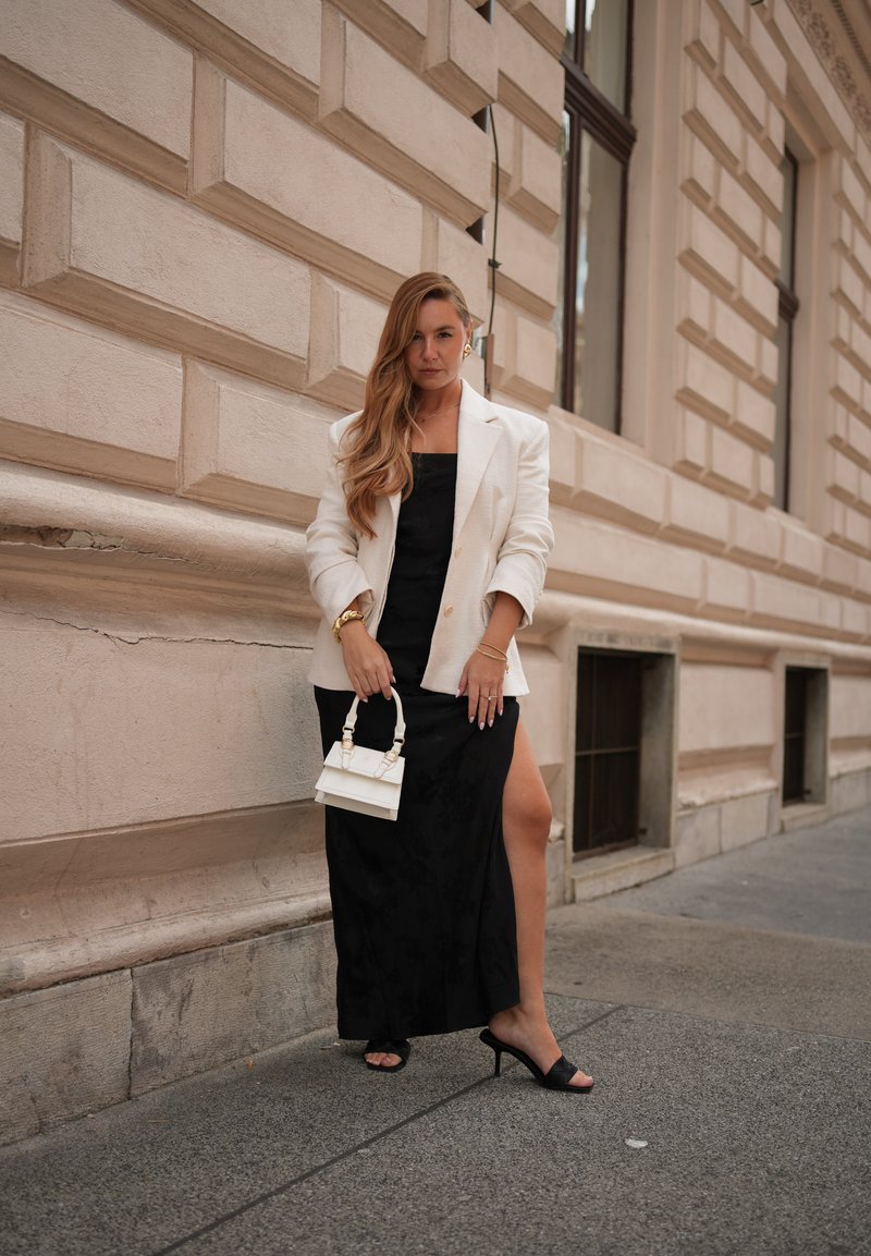Blazer blanc sur une robe maxi fleurie noire avec une fente sur la cuisse. Des mules noires à talons et un petit sac à main blanc complètent le look.