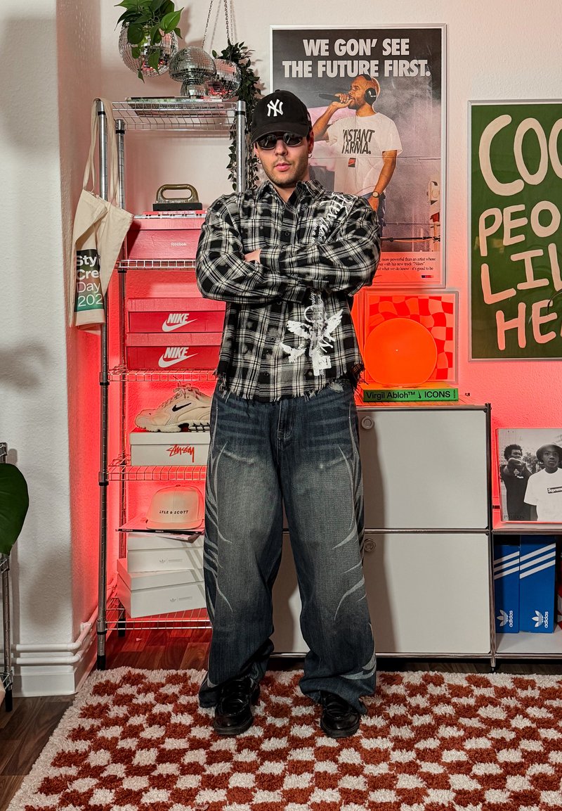 Homme portant des lunettes de soleil, une casquette noire et une chemise à carreaux, debout les bras croisés dans une pièce avec des boîtes de baskets, des affiches et des plantes.