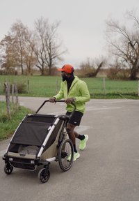 Løpevogn med svart nettoppdekning og beige kropp, dyttet av en person iført en knallgrønn jakke og oransje lue på en landlig sti.