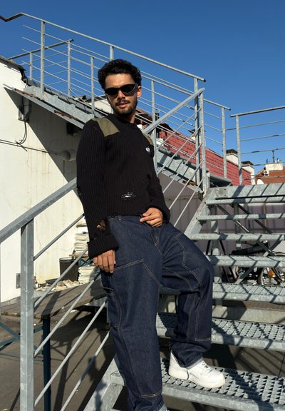 Suéter negro acanalado con detalles verdes y rasgaduras, combinado con jeans de denim azul suelto y zapatillas blancas, sobre escaleras de acero.