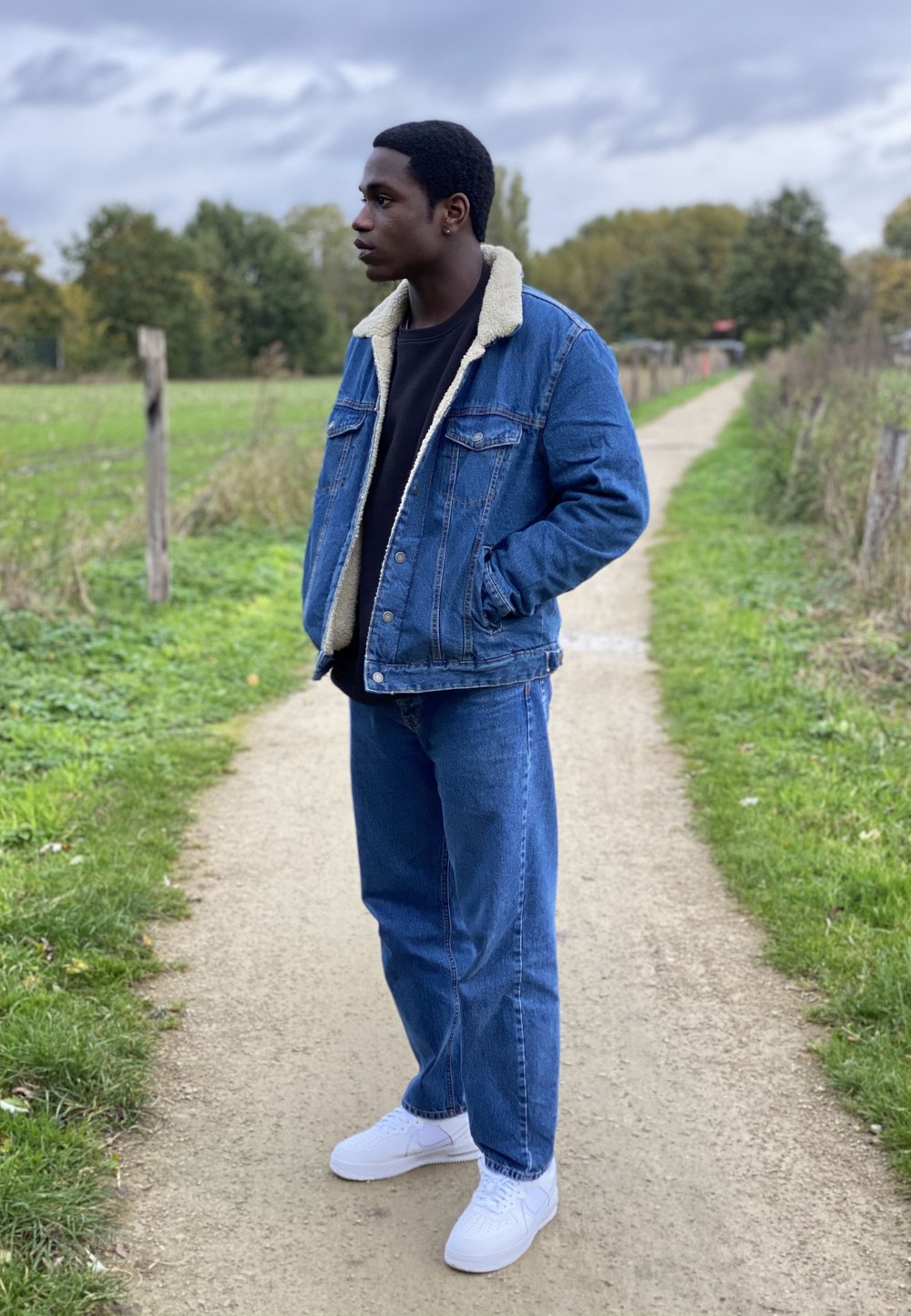 Denim jacket with a shearling collar, black shirt, loose-fitting denim jeans, and white sneakers, standing on a path surrounded by greenery.