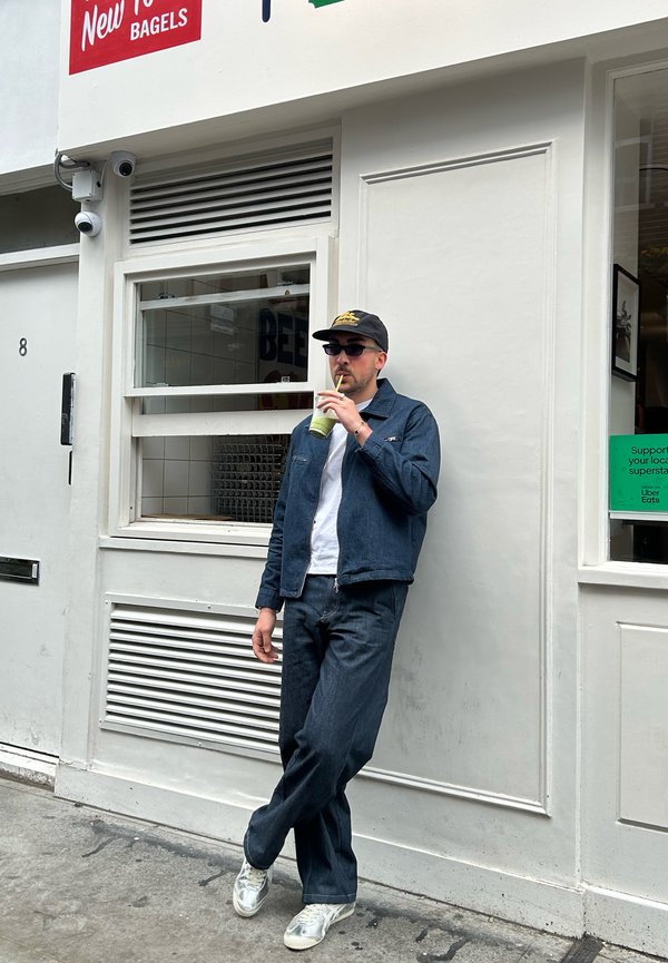 Giacca e pantaloni in denim blu scuro, abbinati a una maglietta bianca e un frullato verde. Appoggiato contro un negozio con una facciata bianca.