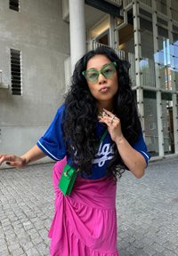Blue sports jersey with white detailing, pink skirt, green sunglasses, and a small green crossbody bag. Curly black hair, posing outdoors.