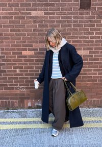 Námořnický dlouhý kabát, béžová šála, pruhované světle modré tričko, široké hnědé kalhoty, šedé tenisky, olivově zelená kabelka, s šálkem kávy v ruce.