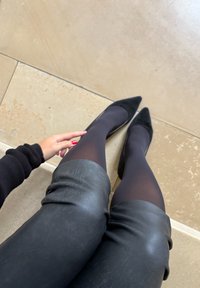 Person som bär svarta tights, spetsiga svarta skor och en svart kjol sitter på ett beige kaklat golv, med en hand vilande nära knäet med röd nagellack.