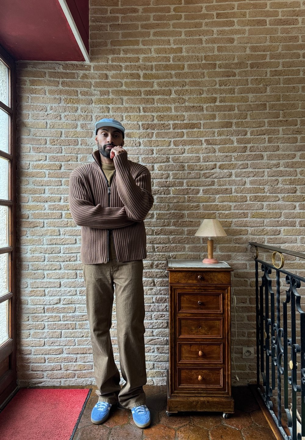 Suéter marrón de canalé con cremallera, camisa olivo, pantalones marrones, zapatillas azul. Mesita de noche de madera con cinco cajones y una pantalla de lámpara color crema. Fondo de pared de ladrillo.