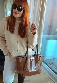 Femme aux cheveux roux portant des lunettes de soleil noires, un cardigan beige, un pantalon blanc, tenant un grand sac à main en cuir marron à texture crocodile à l'intérieur.