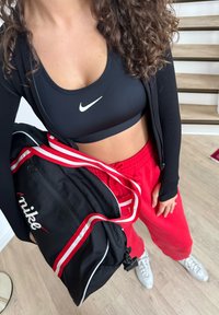 Femme en brassière de sport noire, veste noire, pantalon de survêtement rouge, baskets blanches, tenant un sac de sport Nike noir avec des sangles rouges et blanches sur un sol en bois.