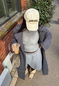 Persona con gorra beige de Diesel, abrigo largo gris, top ajustado gris, falda gris, botas beige abiertas en los dedos, sosteniendo un café helado y un bolso peludo al aire libre.