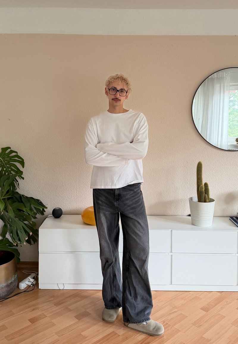 Jeune personne aux cheveux blonds bouclés et portant des lunettes, debout les bras croisés dans une pièce minimaliste avec des plantes et un miroir mural rond.