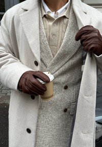 Beige ullkåpe over en teksturert beige vest og en brunaktig strikket polotrøye, mørkebrune lærhansker, holder en kaffekopp med et brunt erme.