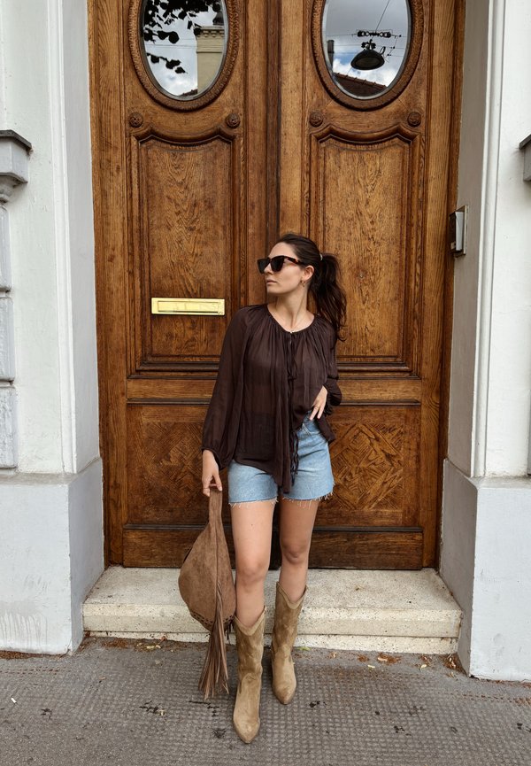 Frau mit Sonnenbrille, brauner Bluse, Jeansshorts und beigen Stiefeln steht vor großen Holztüren und hält eine braune Tasche.