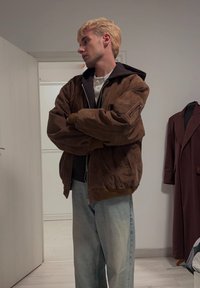 Giacca bomber in suede marrone con cappuccio, tasche con zip e polsini a costine, abbinata a jeans a gamba larga blu chiaro in un ambiente neutro.