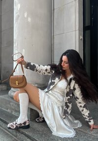 Cowprint denim jacket, white dress with lace trim, knee-high white socks, and cowprint loafers. Brown crossbody bag with gold hardware.