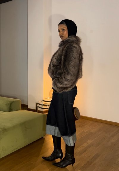 Chaqueta de piel sintética marrón, falda de mezclilla negra con dobladillo en contraste, botas negras de tacón alto y un pañuelo negro sobre un fondo neutro.