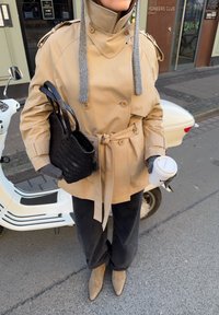 Beżowy płaszcz trencz z paskiem w talii i wysokim kołnierzem, szare dzianinowe dodatki, czarna pleciona torba, ciemne spodnie i beżowe szpiczaste botki.